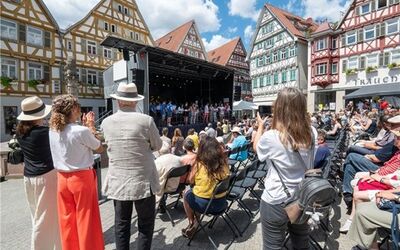 Auf dem Herrenberger Marktplatz. Im Vordergrund stehen 4 Personen. Das Publikum in den Stuhlreihen applaudiert dem Riesenorchester auf der Bühne.