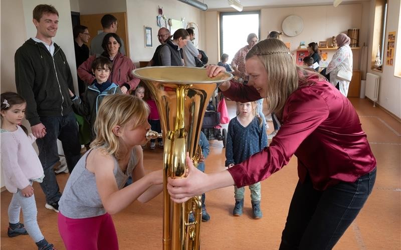 Ein mit vielen Personen gefüllter Raum der Musikschule. Im Vordergrund hält die Tubalehrerin eine Tuba für ein Kind fest, das in die Tuba bläst.