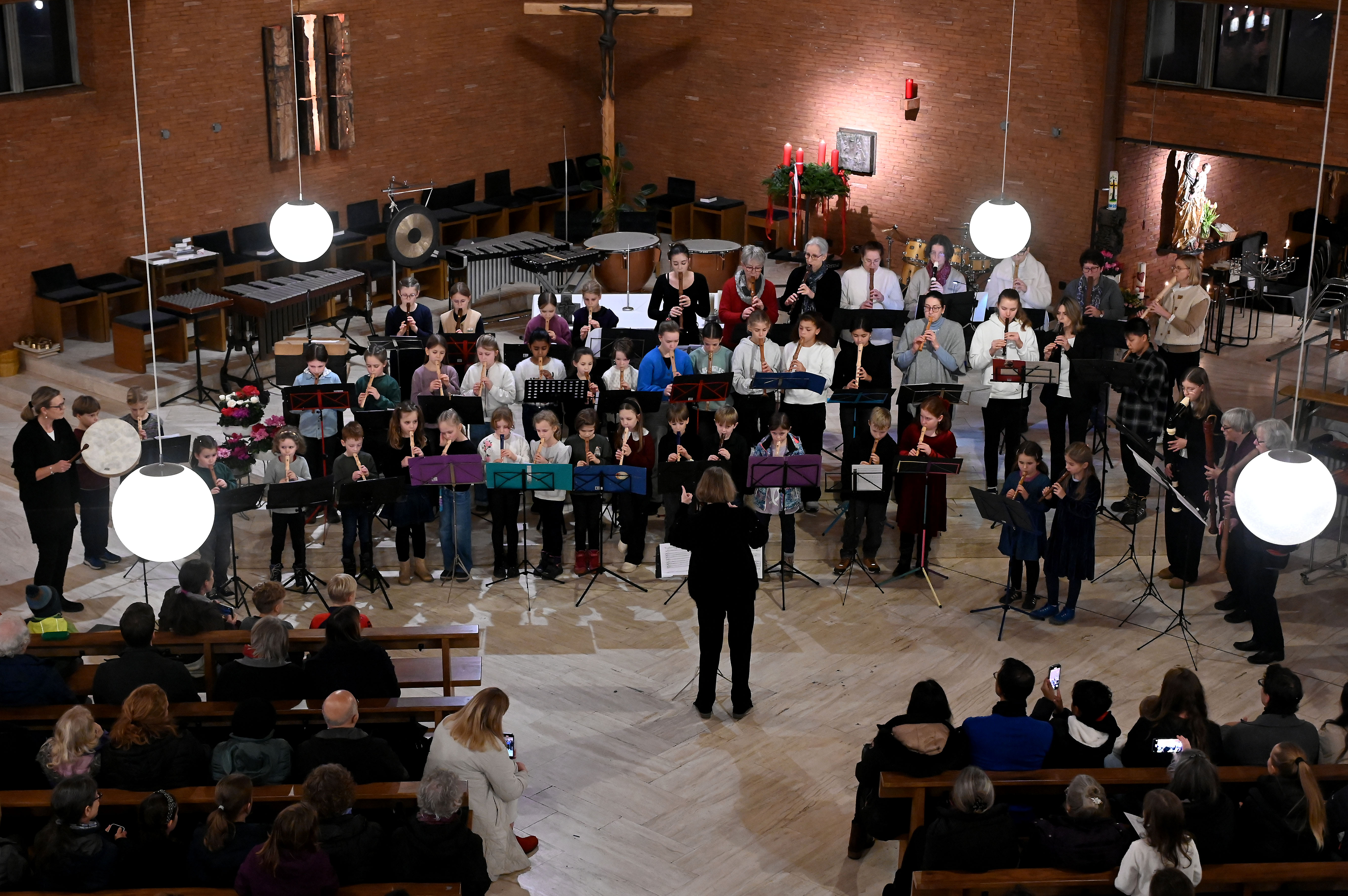 Eine Gruppe von Kindern spielt Blockflöten in einem Kirchenkonzert, dirigiert von einer Person, während das Publikum zuschaut.